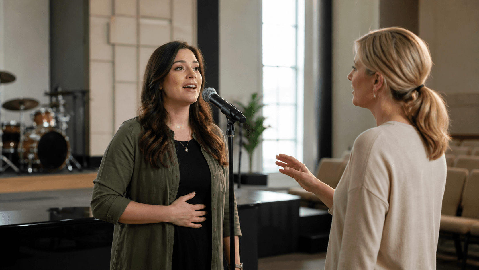 Worship leader sings into a microphone under warm stage lights, with a calm coach nearby in soft focus.